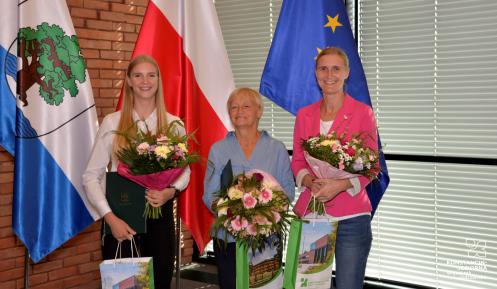 Trzy osoby stoją obok siebie w rzędzie – młoda dziewczyna z długimi blond włosami, kobieta w średnim wieku oraz młoda kobieta. Każda w rękach trzyma bukiet kolorowych kwiatów i torbę prezentową ze zdjęciem Urzędu Miasta i napisem Konstancin-Jeziorna.
