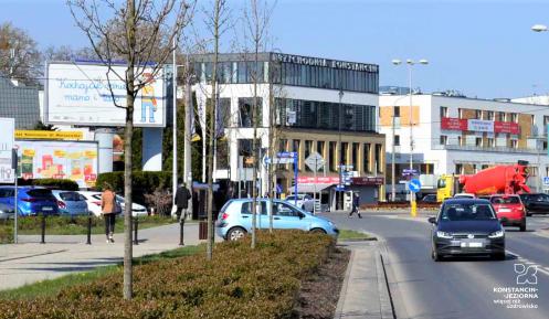 Ulica dwukierunkowa w Konstancinie-Jeziornie. Po ulicy jadą samochody, po chodnikach idą piesi. W dali widać budynki oraz duże banery reklamowe. 