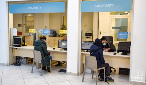 Urząd, dwie osoby siedzą na krzesłach przed stanowiskami paszportowymi.