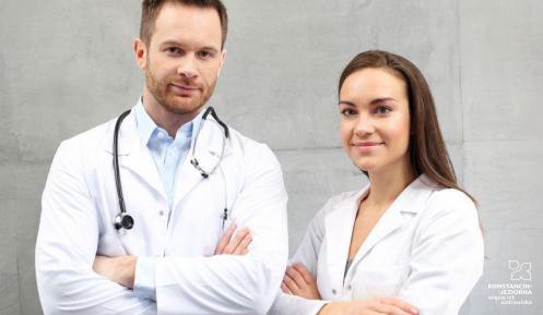 Po lewej stronie stojący mężczyzna w białym fartuchu lekarskim. Na szyi ma zawieszone słuchawki. Po prawej stronie kobieta w białym fartuchu. Para lekarzy stoi na tle z betonu.