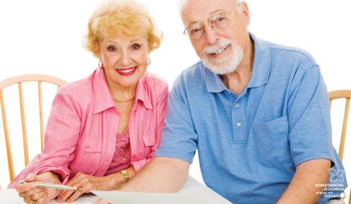 Starsi mężczyzna i kobieta siedzą na krzesłach i uśmiechają się, W dłoniach trzymają kartki.