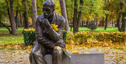 Marmurowy pomnik Stefana Żeromskiego, który siedzi na ławce. Ma zamyśloną minę, lewą ręką podpiera głowę, prawą ma położoną na kolanie. W tle wysokie drzewa, na trawie leżą jesienne liście. 