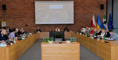 Posiedzenie członków Rady Miejskiej oraz władz gminy. Osoby siedzą przy stołach ułożonych na kształt podkowy. Na środku sali stoi duży telewizor, a na ścianie wisi biały ekran. 