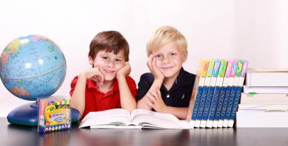 Dwóch chłopców (blondyn i szatyn) siedzi uśmiechniętych przy biurku. Na biurku znajdują się: globus, podręczniki, atlasy i duzy globus.