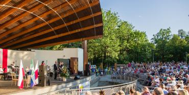 Zdjęcie przedstawia amfiteatr w Parku Zdrojowy w letni dzień. Na scenie występują muzycy. Widownia wypełniona jest w całości. W dali widać wysokie drzewa. 