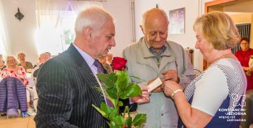 Sala. Trzy osoby w średnim wieku, dwóch mężczyzn i kobieta, stoją obok siebie. Jeden z mężczyzn w ręku trzyma mikrofon i czerwoną różę.