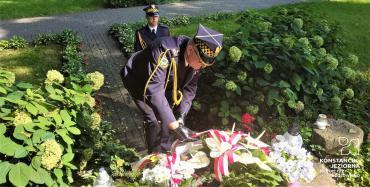  Dwóch strażników miejskich, ubranych w stroje galowe, składa biało czerwone kwiaty pod  pomnikiem upamiętniającego bohaterów Bitwy Warszawskiej z 1920 roku. 