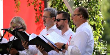 Czterech mężczyzn – śpiewaków, stoi obok siebie w rzędzie. Ubrani są w białe koszule. W rękach trzymają śpiewniki. 