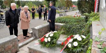 Cmentarz. Para emerytów stoi przed pomnikiem ofiar drugiej wojny światowej. Przy pomniku stoi mężczyzna ubrany w mundur Straży Miejskiej. Na pomniku leżą dwa wieńce z biało-czerwonymi kwiatami.