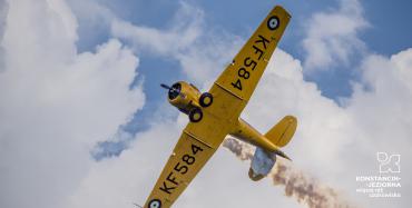 Na błękitnym niebie z chmurami leci żółty zabytkowy samolot. Na skrzydłach są czarne napisy z nazwą modelu – KF584. Za samolotem ciągnie się dym.