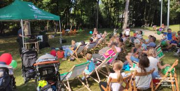 Park Zdrojowy. Po lewej stronie pod zielonym namiotem stoją dwie osoby. Naprzeciwko nich siedzi na krzesłach i na leżakach grupa około trzydziestu osób – dzieci i osoby dorosłe.