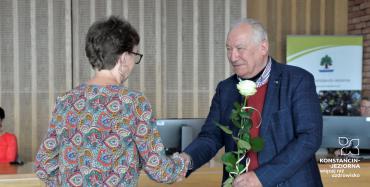 Sala konferencyjna. Mężczyzna w średnim wieku, ubrany w garnitur naprzeciwko niego stoi kobieta, której wręcza białą różę.