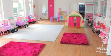 Wnętrze dużej sali z ustawionymi pod ścianami zabawkami dla dzieci, na podłodze leżą dywaniki w kolorach różowym i białym, po lewej i prawej stronie okna w ścianach, na wprost widoczne przez otwarte drzwi łazienka