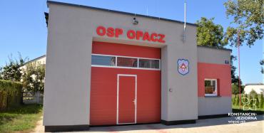 Duży budynek widziany od frontu, pośrodku duża brama wjazdowa w kolorze czerwony, nad nią napis OSP OPACZ w kolorze czerwonym