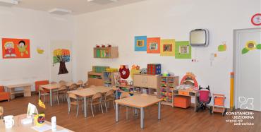 Duża sala z jasnmi ścianami, pośrodku stoją trzy małe stoliki i krzesełeczka dla dzieci, pod ścianą po prawej stroni stoją meble dziecięce wypełnione zabawkami, na ścianach kolorowe i wesołe obrazki