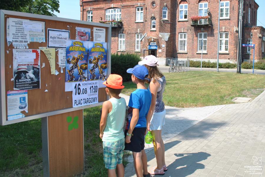Troje dzieci czytających ogłoszenia umieszczone na brązowej tablicy informacyjnej, w tle budynek z czerwonej cegły.