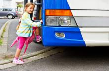 Dziewczynka ubrana w różową sukienkę i zielony sweter wsiada do biało-niebieskiego autobusu.