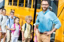 Młody mężczyzna na tle żółtego autobusu szkolnego oraz wsiadających do niego dzieci.