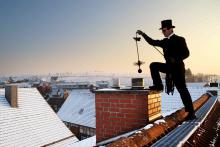 Mężczyzna, kominiarz stoi na dachu. Ubrany jest w czarną kurtkę, na głowie ma czarny kapelusz. Czysci komin. W tle domy jednorodzinne i błękitne niebo.