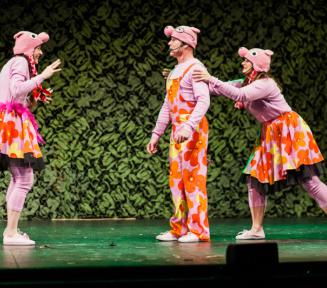 Plakat promujący premierę spektaklu. Trzy osoby stoją obok siebie na scenie. Mężczyzna i dwie kobiety. Ubrani są w pomarańczowo-różowe stroje. Na głowach mają różowe czapki przedstawiające głowę świnki.