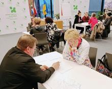 Mieszkańcy uczestniczą w warsztatach konsultacyjnych w sali urzędu, którzy przy stolikach omawiają mapy i dokumenty z przedstawicielami gminy.