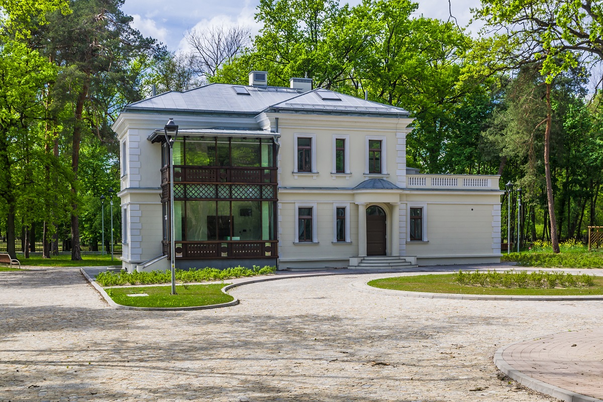 Piętrowy budynek willowy, z lewej drewniana piętrowa weranda, po prawej niska przybudówka, przed budynkiem zagospodarowany teren, z tyłu duże drzewa