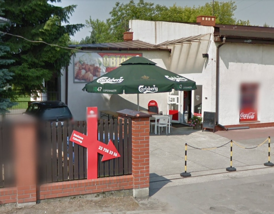 Na pierwszym planie widoczne ogrodzenie, za którym dobudowany obiekt do większego budynku, przed nim parasol i stoliki, po parwej widoczne drzewa