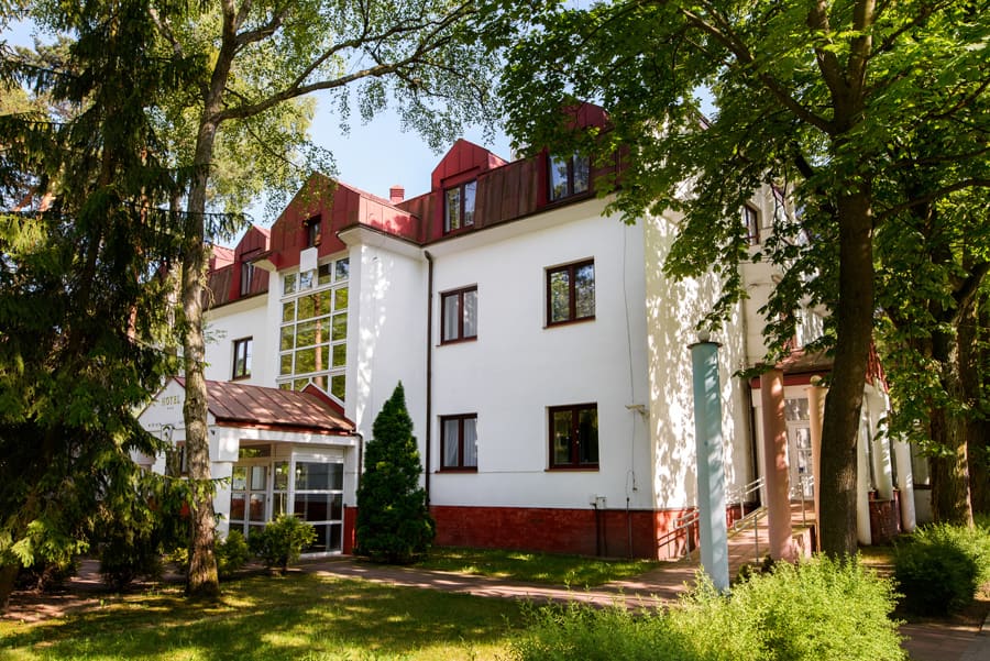 Duży obieky dwukondygnacyjny z wysokim poddaszem z oknami, ściany białe, dach w kolorze czerwonym, wejście z dodanym oszkleniem i zadaszeniem, wokół obiektu drzewa
