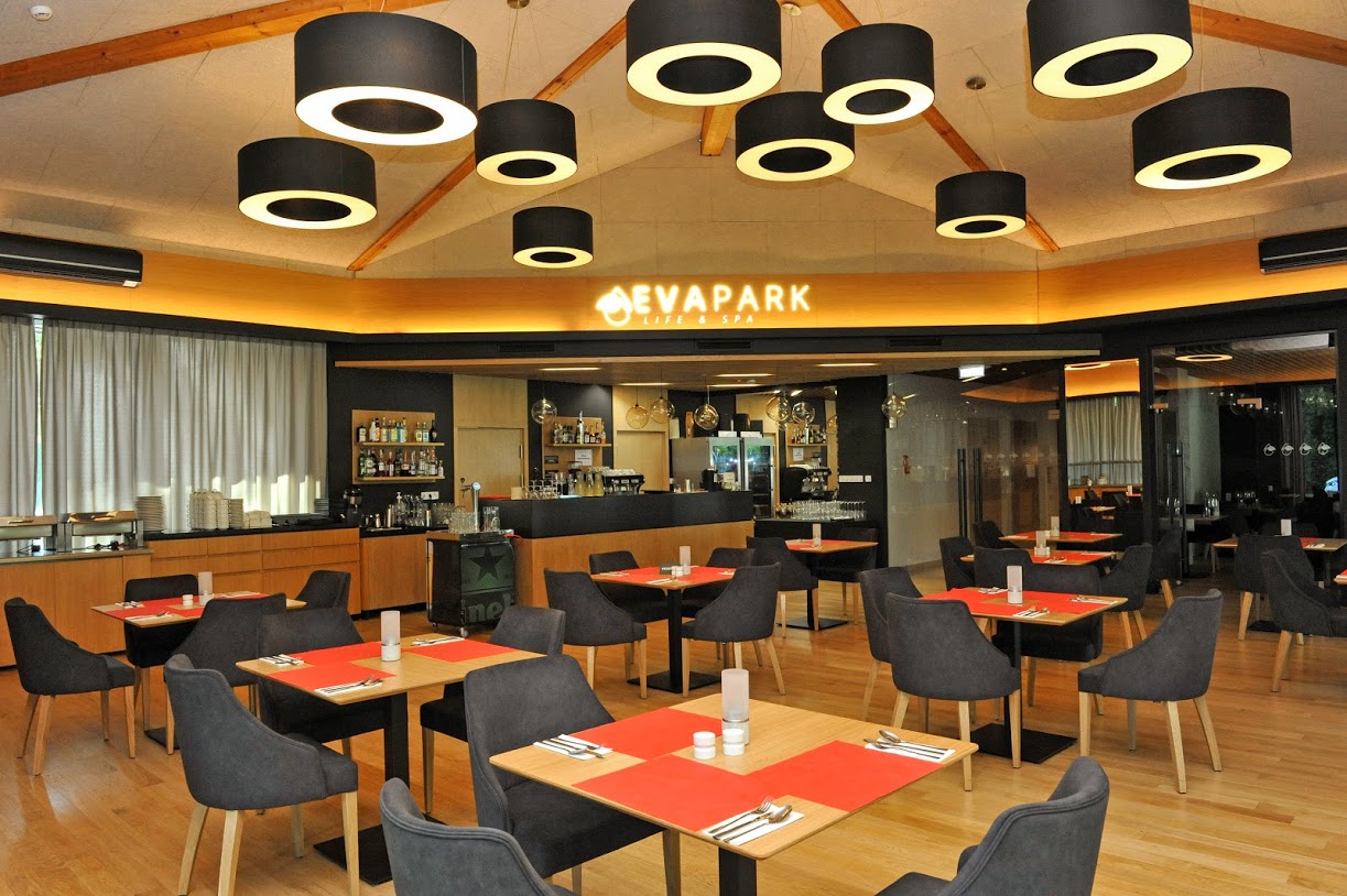 Wnetrze nowoczesnej restauracji, pośrodku nakryte stoliki z fotelikami, naśrodku i po lewej stronie widoczny bar z zastawą, po prawej przeszkolne dzrwi wejściowe, spod sufitu zwisają koliest lampy, pośrodku nad barem neon z nazwalokalu