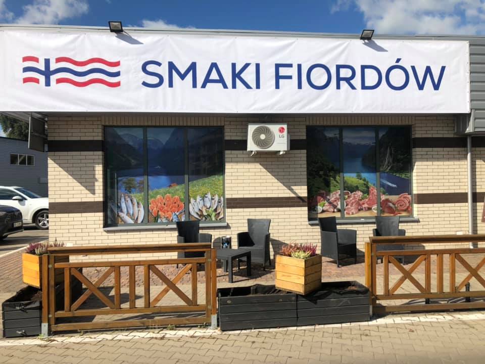 Fragment niskiego budynku, od góry pod dachem baner z nazwaSmaiki Fiordów i znakiem graficznym, ściana z duzymi oknami z rysunkami ryb, przed budynkiem fotele i stolik, oraz niskie ogrodzenie