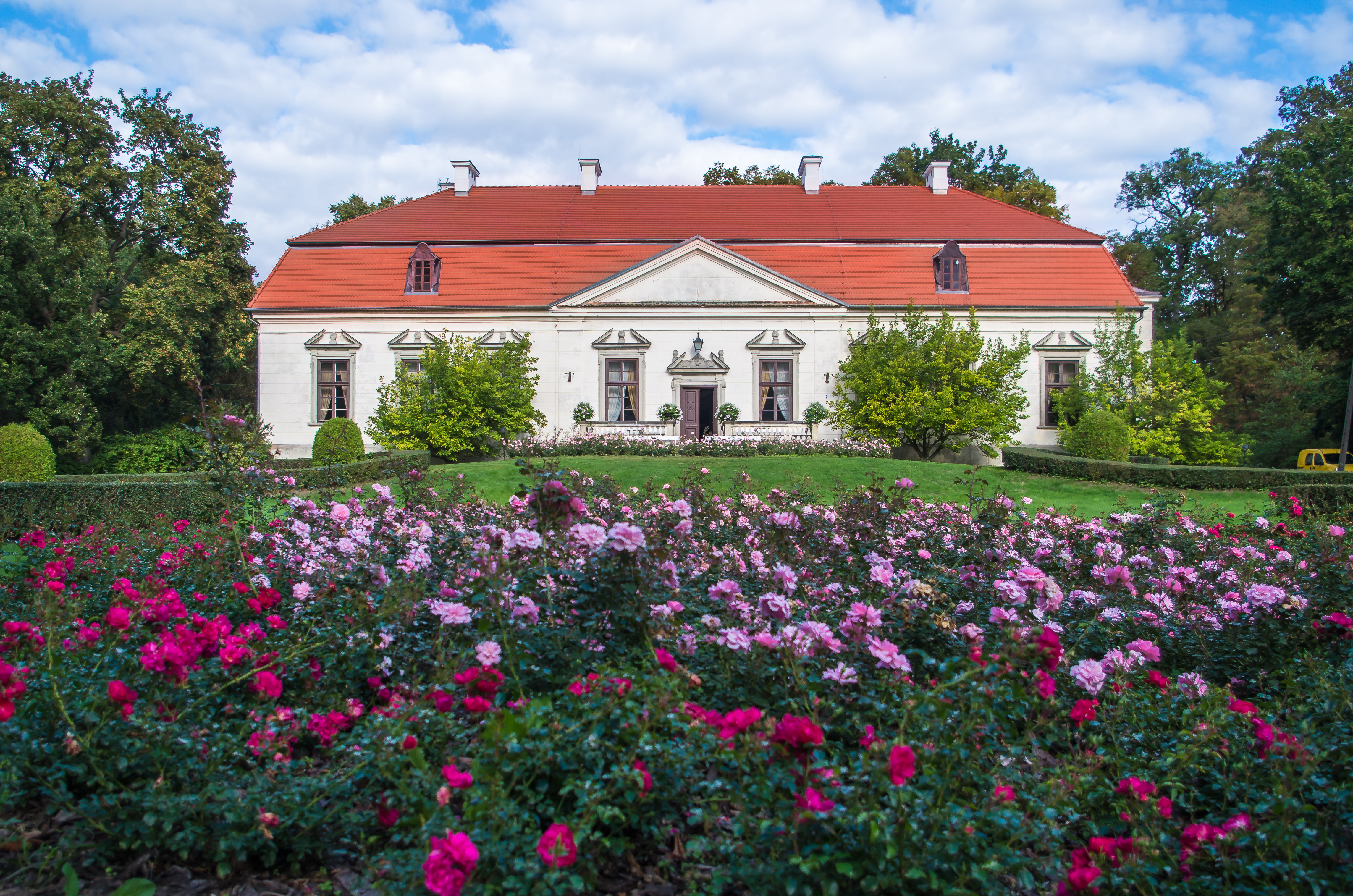 Widok na niski dwór z pomarańczowym dwuspadowym dachem. Przed budynkiem znajduje się duża rabata z fioletowych kwiatów.