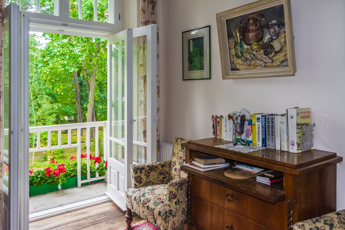 Wnętrze willi ze stylizowanymi meblami oraz książkami i obrazami. Widok z otwartego na balkon okna rozpościera się na ogród 