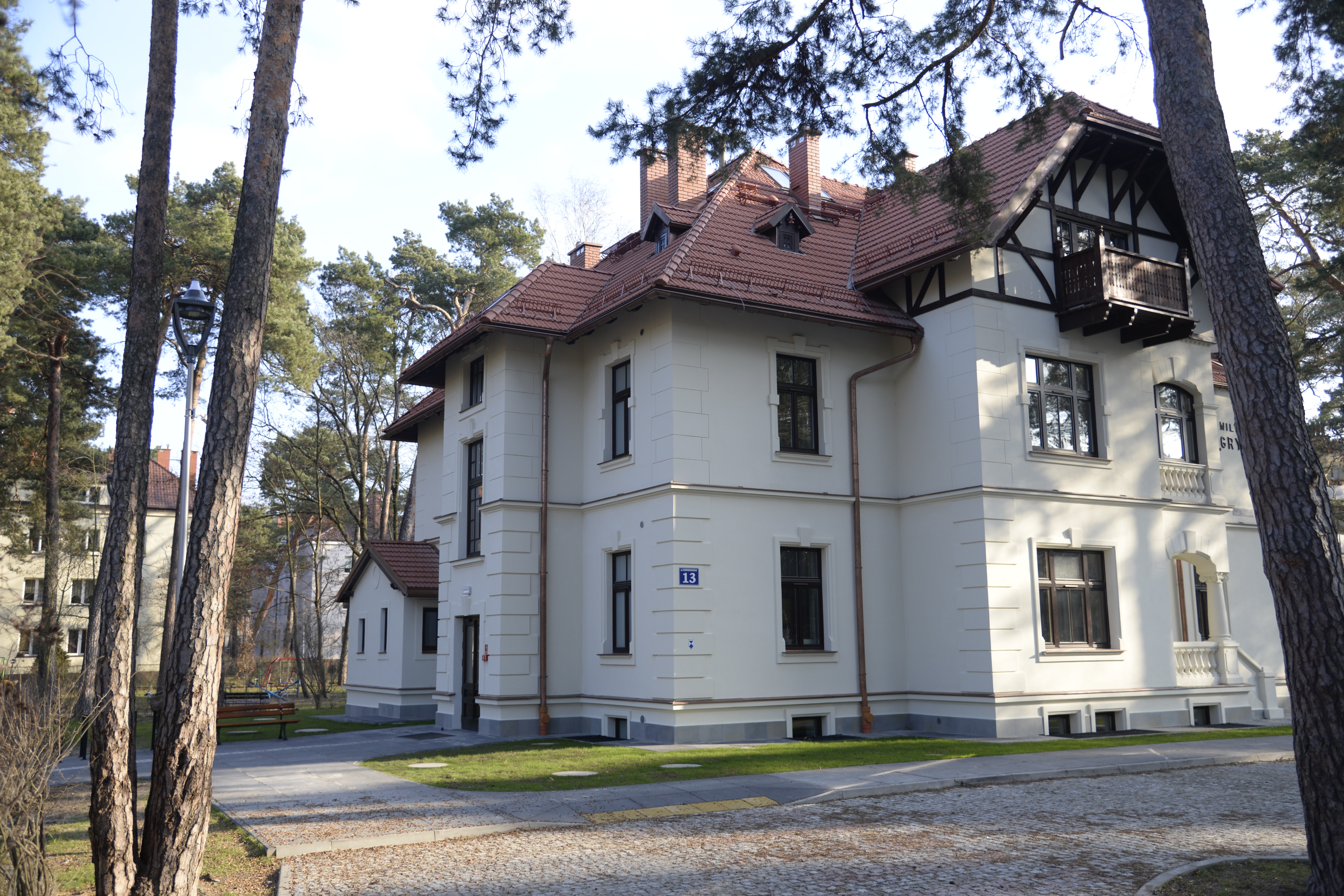 Widok na willę Gryf od strony pobliskiego placu zabaw. Odrestaurowany budynek z jasną elewacją i wielospadowym dachem. W pobliżu rosna wysokie sosny