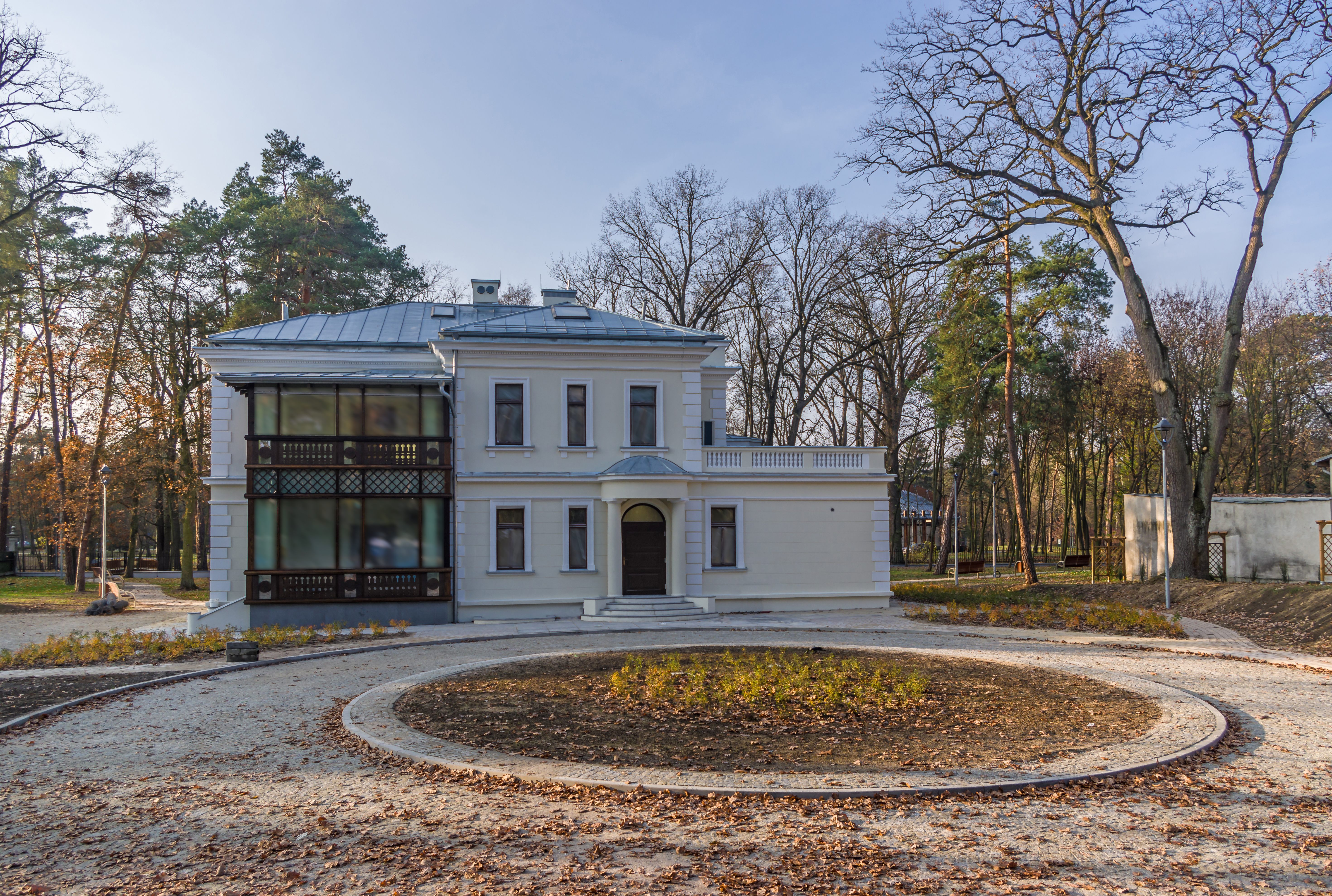 Widok na odrestaurowany budynek willi Kamilin od ulicy Piłsudskiego, z ozdobną drewnianą werandą. Przed budynkiem znajduje się ciąg komunikacyjny z dużym rondem przeznaczonym na nasadzenia ozdobne
