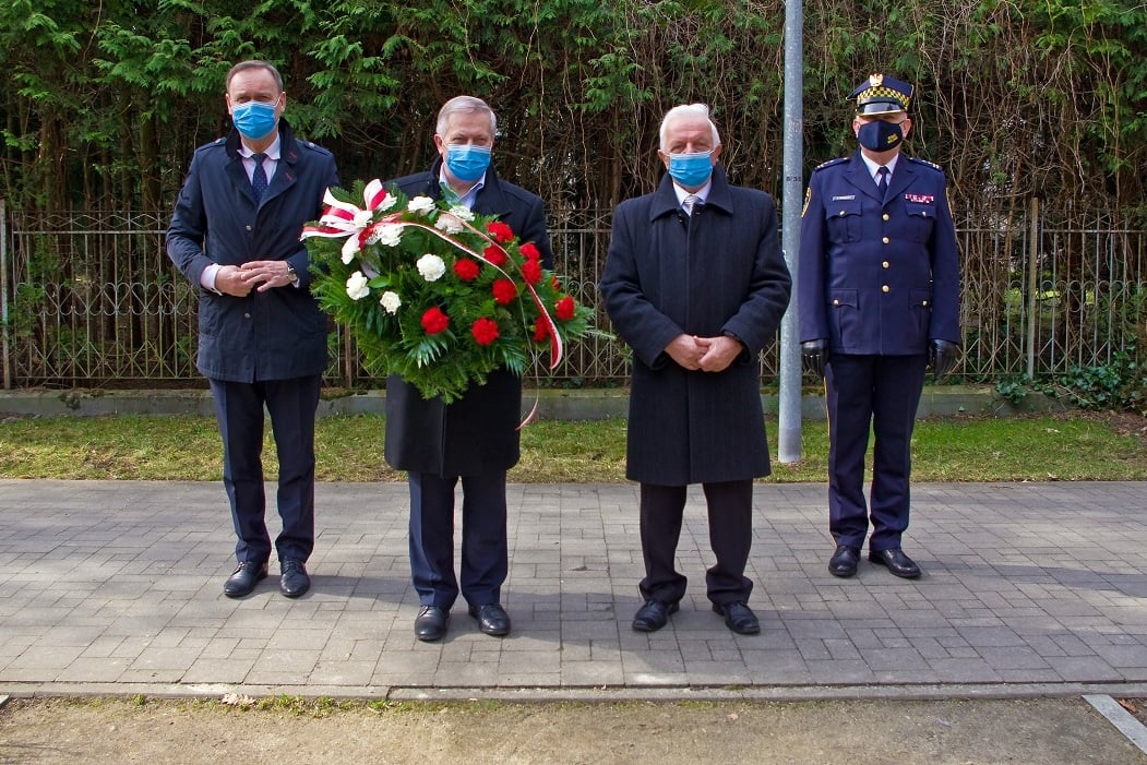Czterej mężczyźni stojący, każdy w maseczce na twarzy, jeden trzyma wieniec biało-czerwony, jeden w mundurze straży miejskiej