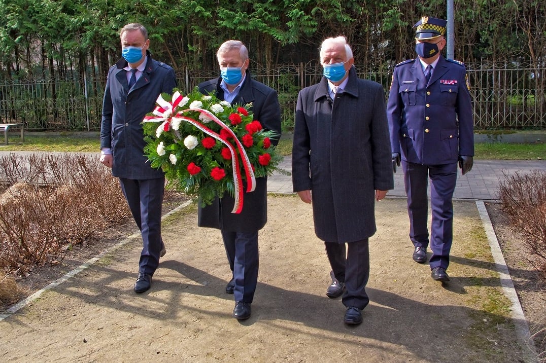 Czterej mężczyźni idą, każdy w maseczce na twarzy, jeden trzyma wieniec biało-czerwony, jeden w mundurze straży miejskiej
