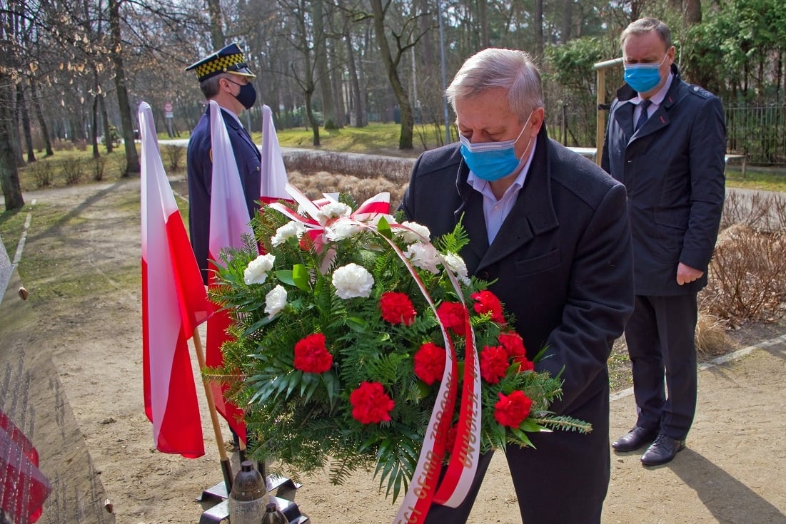Mężczyzna składa wieniec, za nim widoczny inny stojący, obok flagi polska w stojaku