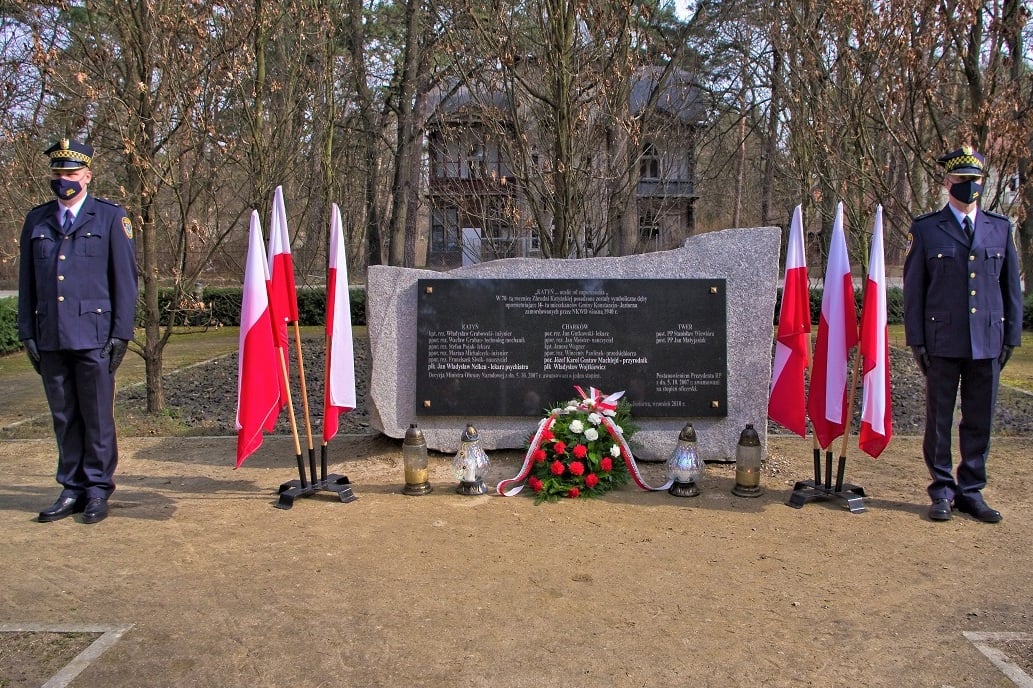 Pośrodku poziomy pomnik kamienny z czarną tablica z tekstem, po bokach flagi polski w stojakach oraz dwaj mężczyźni w mundurach straży miejskiej