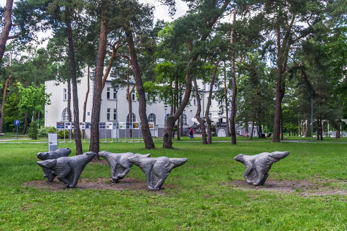 Trzy duże, metalowe rzeźby, które mają postać biegnących wilków. Rzeźby ustawione są na trawie, wśród wysokich drzew. W tle widać wille Hugonówkę
