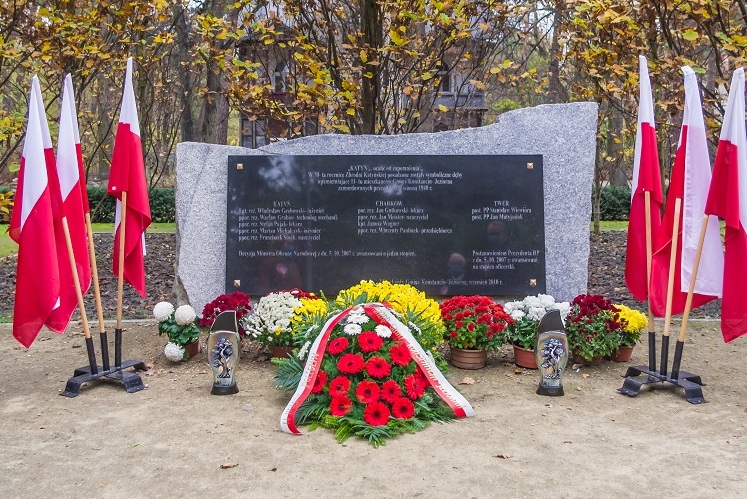 Pośrodku poziomy pomnik kamienny z czarną tablica z tekstem, po bokach flagi polski w stojakach