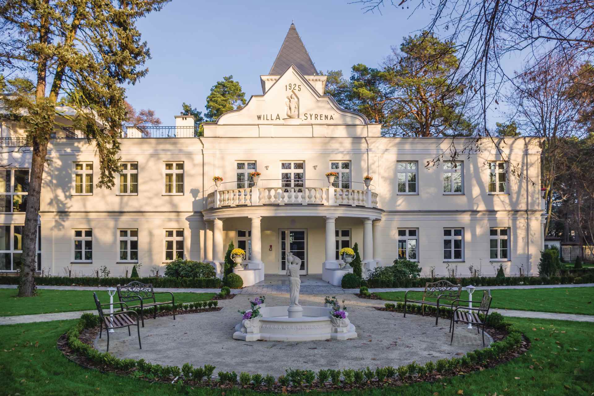 Na pierwszym planie placyk w kształcie okręgu z czterema ławkami i fontanną na środku, w tle budynek z oknami, wieżyczkami oraz balkonem, na szczycie którego znajduje się napis 1925 i willa Syrena.