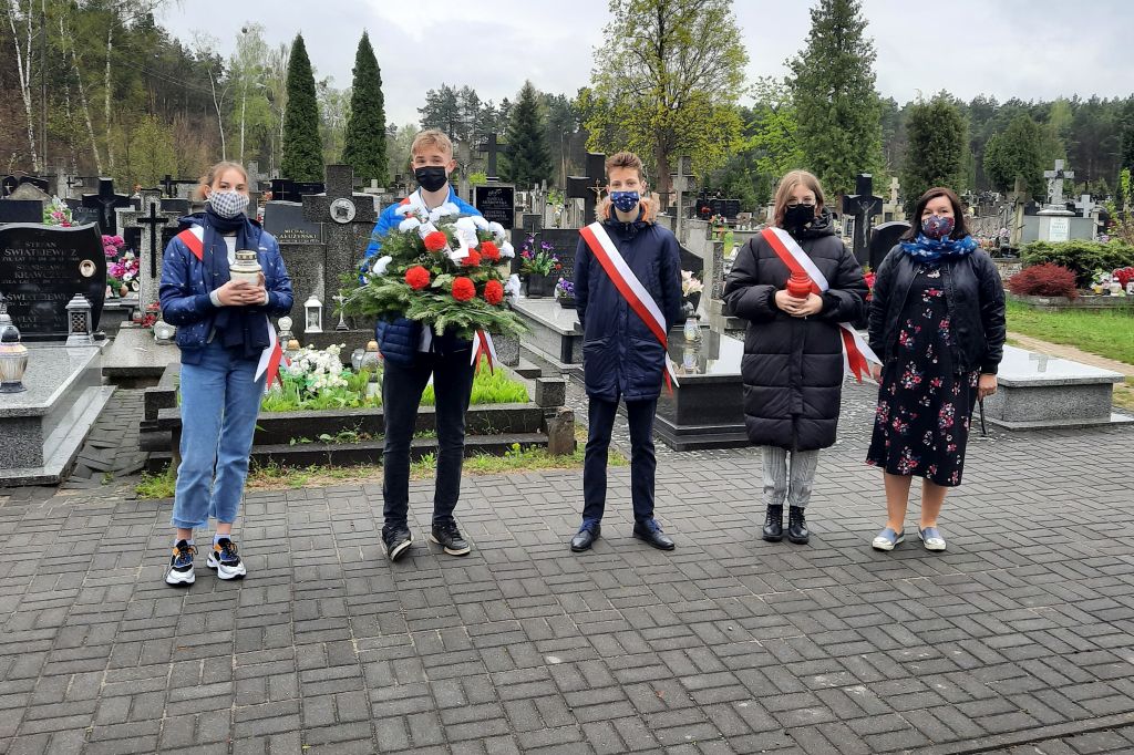 Pięc osób stojących na cmentarzu obok siebie. Dwie osoby trzymają znicz, jedna biało-czerwoną wiązankę z kwiatów . W tle las.