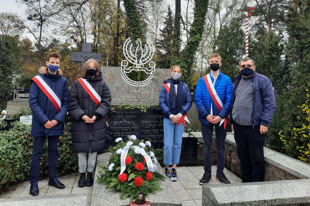 Pięc osób stojących przy pominku kamiennym. Czterech uczniów ma biało-czerwoną szarfę. W tle nagrobki oraz wysokie drzewa.