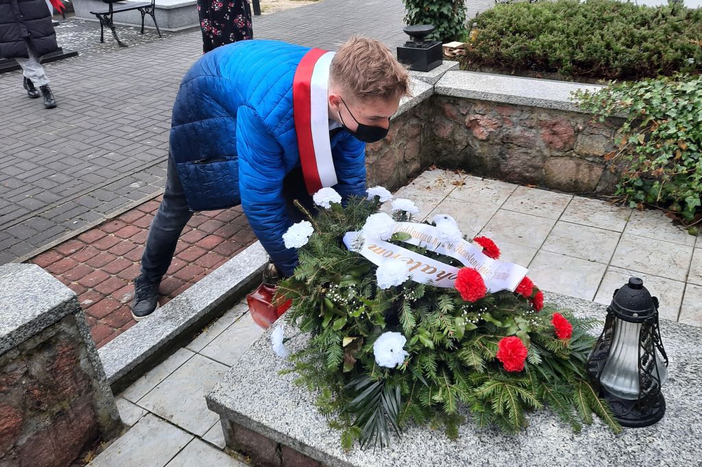 Uczeń w niebieskiej kurtce pochylony nad pominkiem, składa biało-czerwoną wiązankę z kwiatów. 