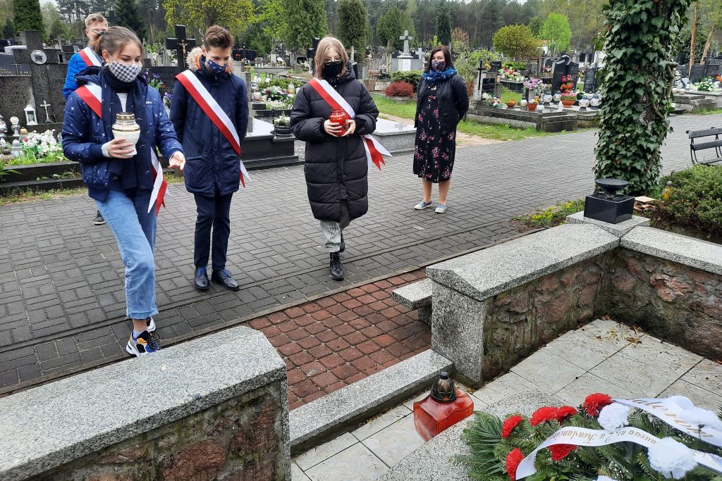 dwie dziewczyny trzymające znicz oraz chłopak w pobliżu pomika kamiennego. w tle nagrobki oraz drzewa.
