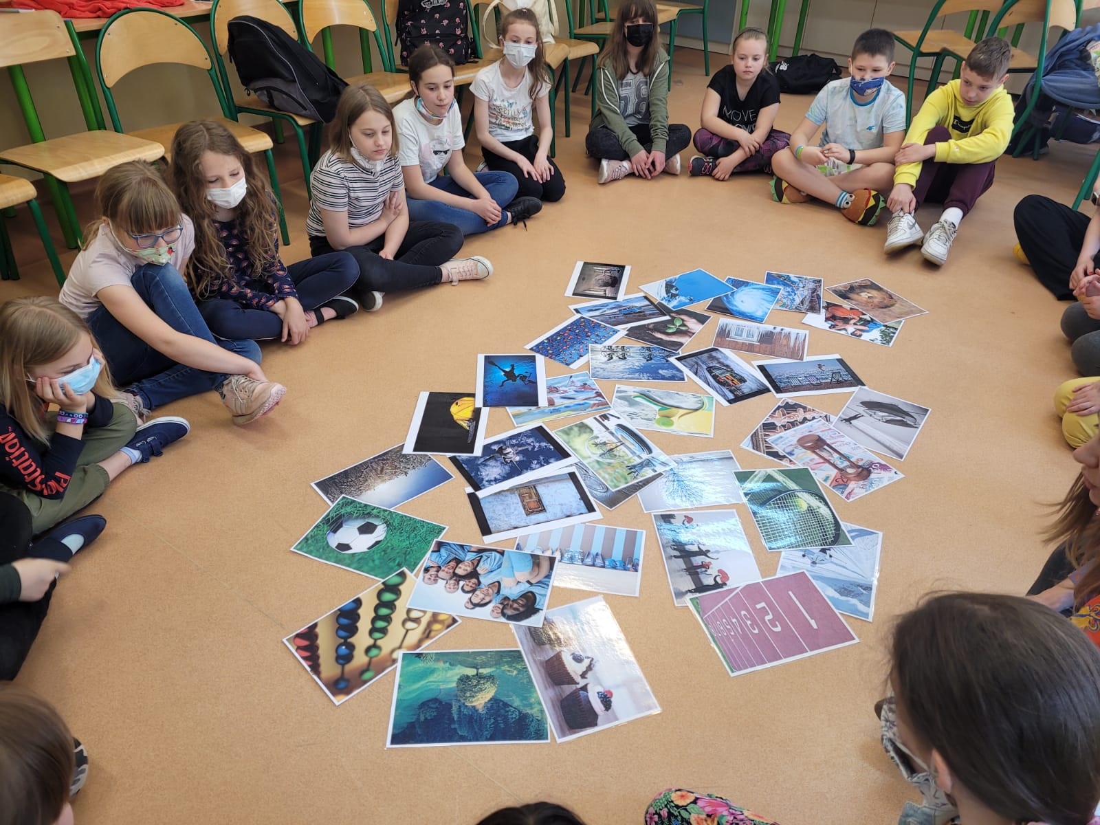 Dzieci siedzące w okręgu na podłodze sali lekcyjnej, spoglądają na rozsypane wewnątrz kolorowe fotografie.
