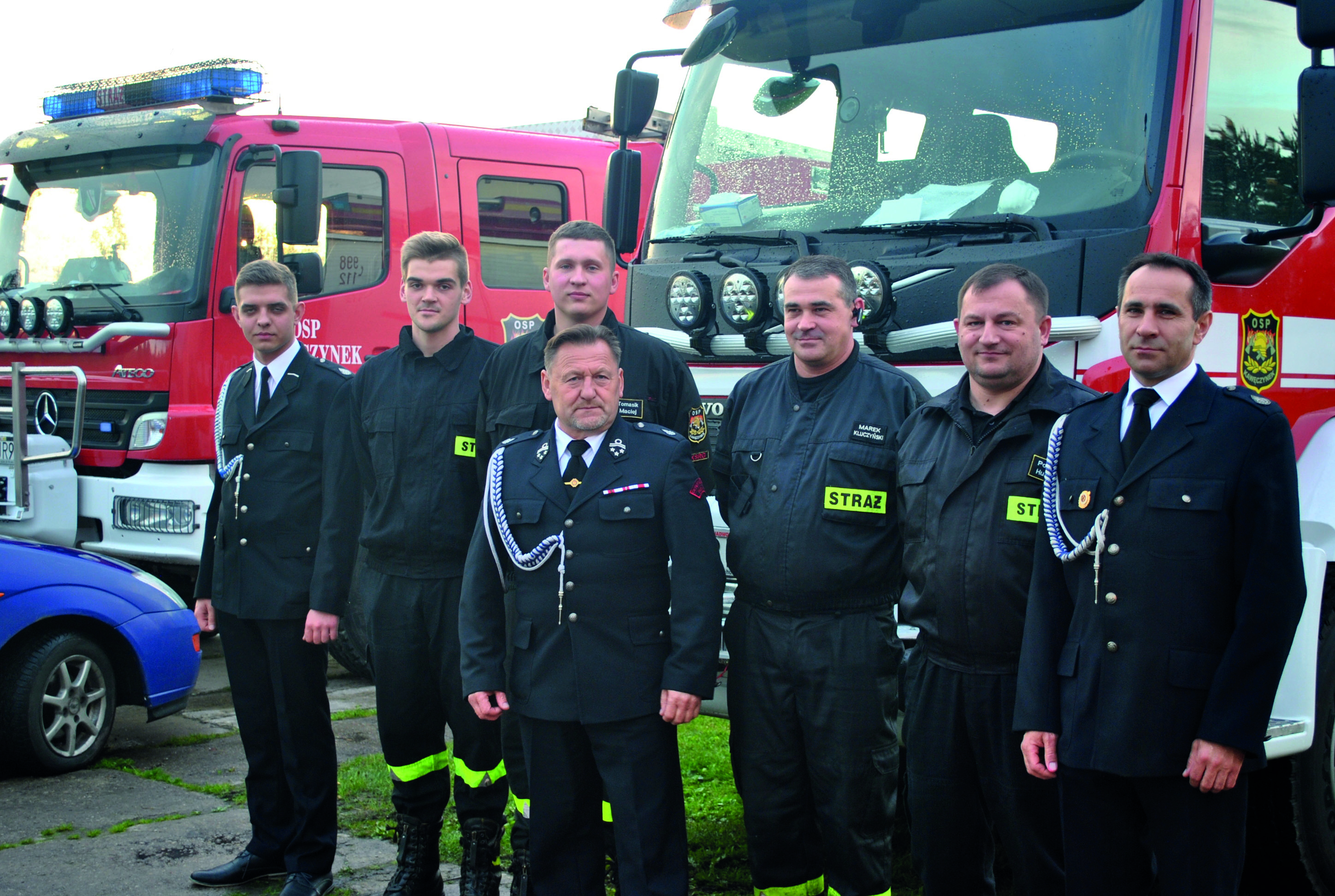 Siedmiu mężczyzn stoi w mundurach strażackich na tle wozów strażackich.