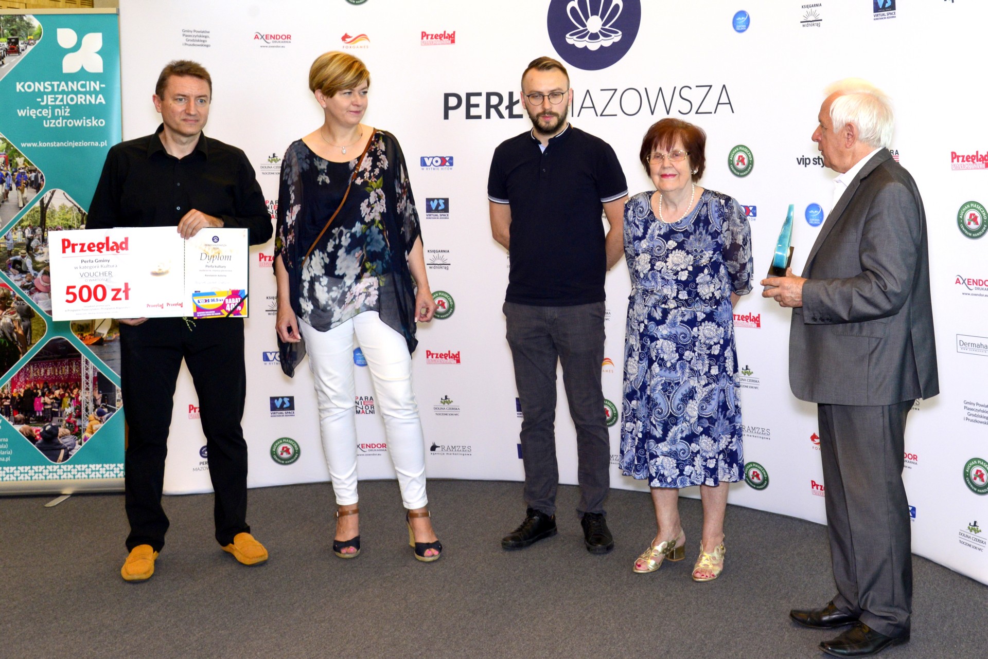 Pięć osób, w tym trzech mężczyzn, z których jeden trzyma dyplom i voucher, a drugi – burmistrz gminy – statuetkę w kształcie perły. W tle ścianka reklamowa.
