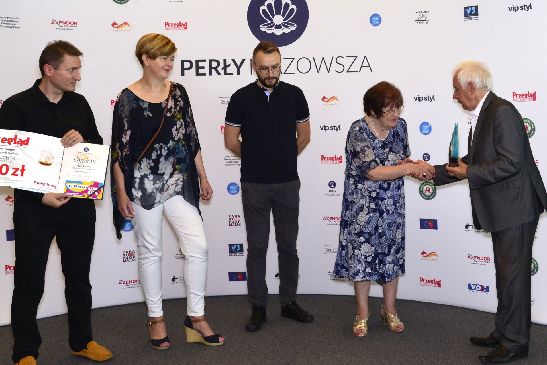 Pięć osób, w tym trzech mężczyzn, z których jeden trzyma dyplom i voucher, a drugi – burmistrz gminy – przekazuje jednej z kobiet statuetkę w kształcie perły i ściska jej dłoń. W tle ścianka reklamowa.