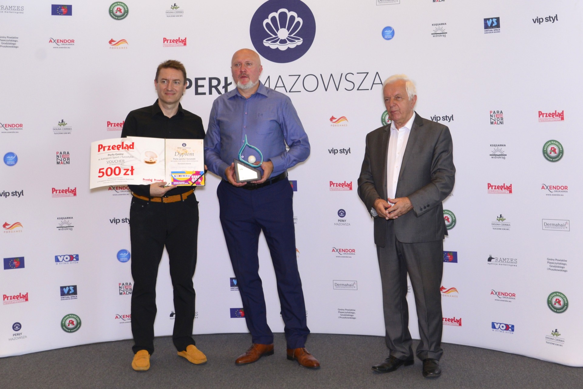 Trzech mężczyzn, z których jeden trzyma dyplom i voucher, a drugi statuetkę w kształcie perły. W tle ścianka reklamowa.